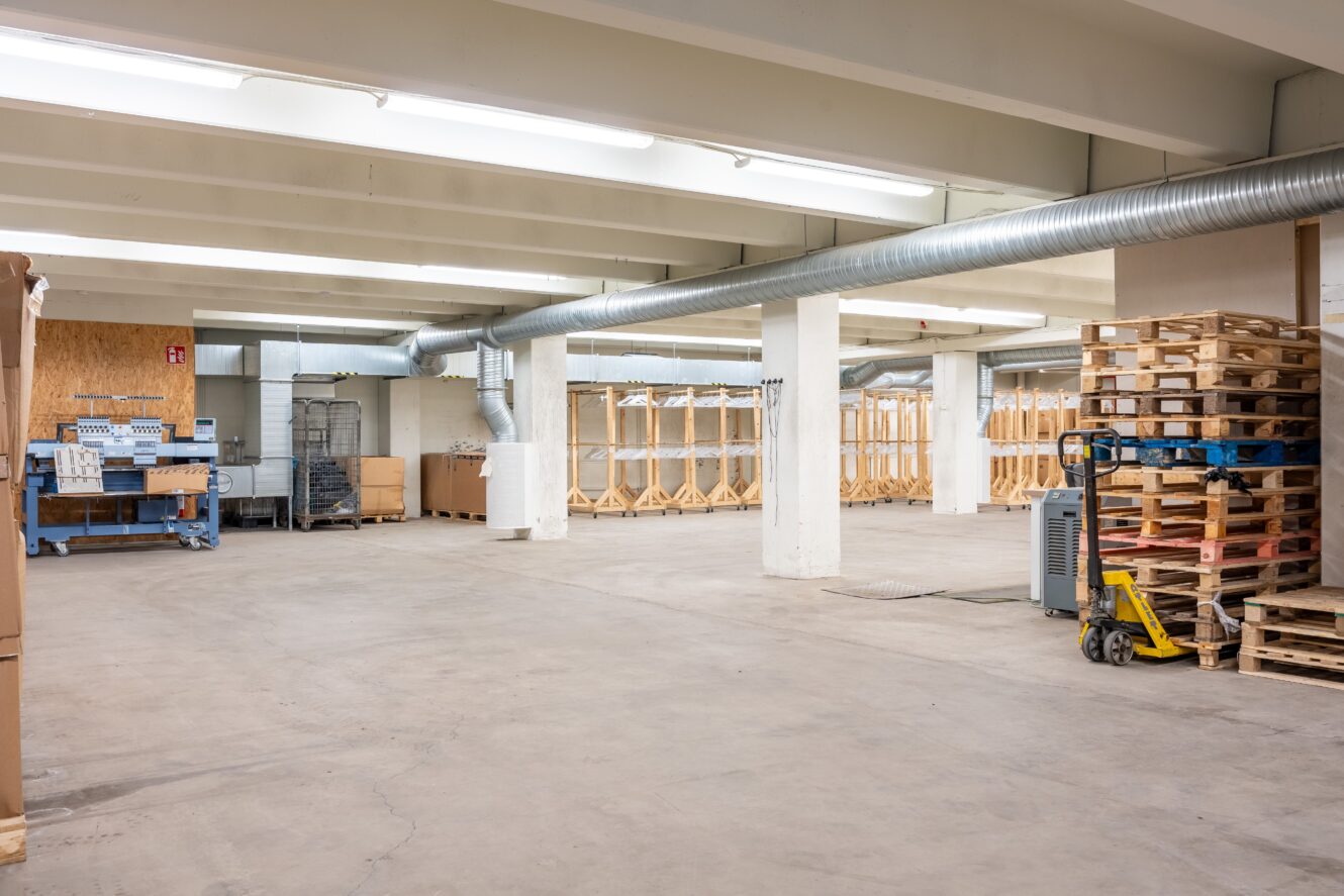 Rymlig lagerlokal med träpallar, lagerhyllor, förpackningsmaterial och industriella ventilationskanaler.