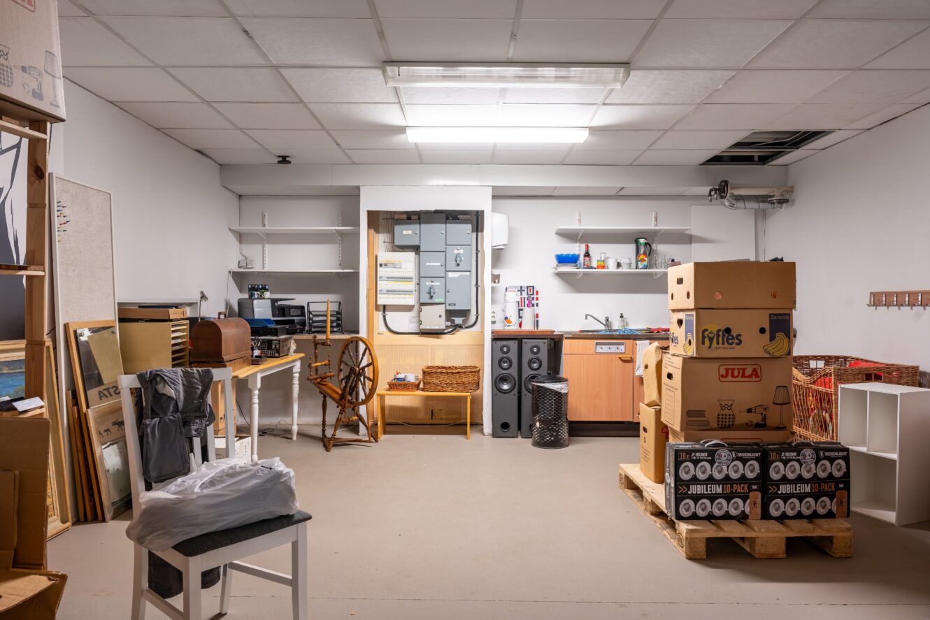 En tvättstuga med lådor på pallar, ett handfat, hyllor, elpaneler, en stol och olika hushållsartiklar längs väggarna under lysrörsbelysning.