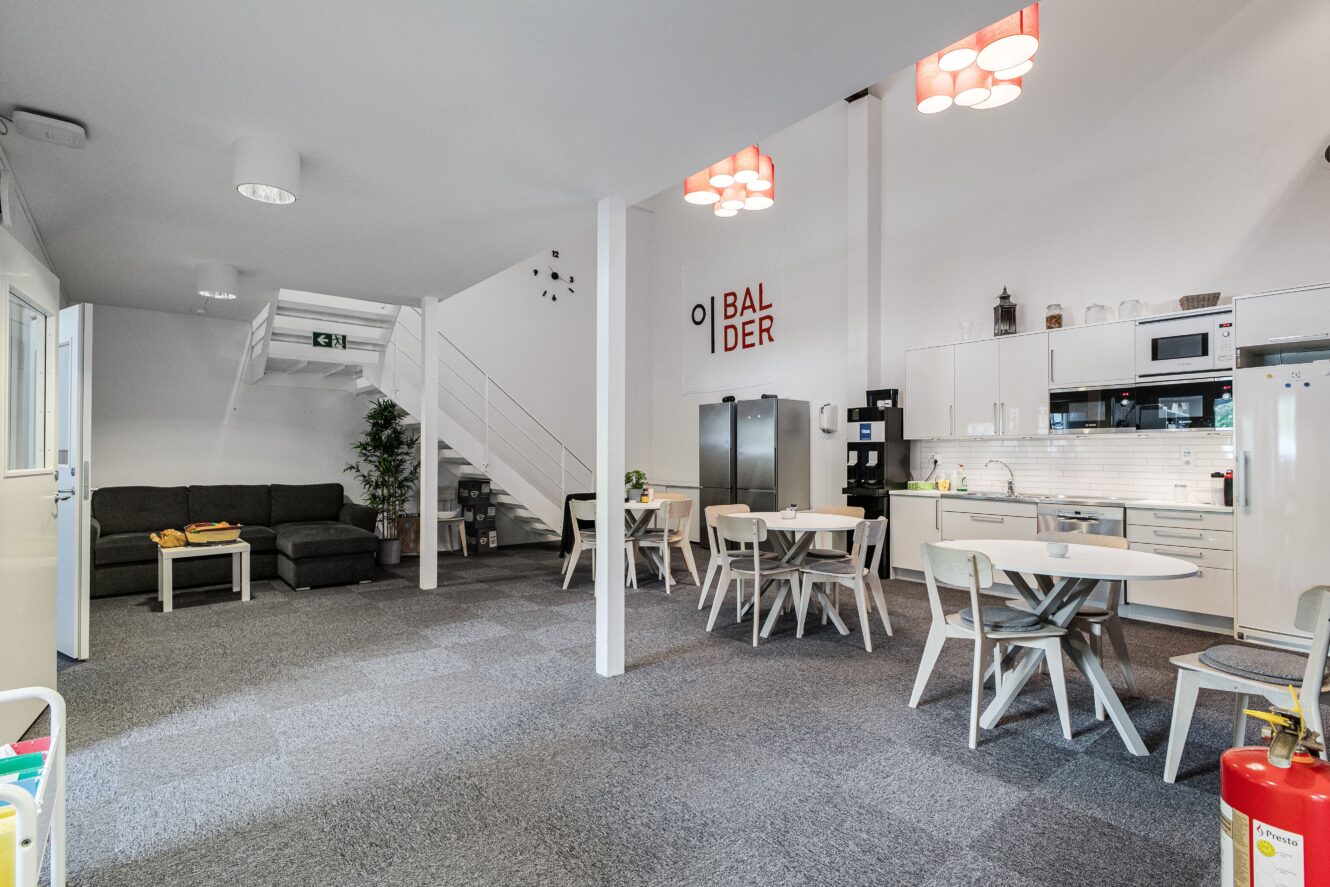 Modernt kontorskök och loungeområde med bord, stolar, soffor, köksmaskiner och trappor i ett rymligt, väl upplyst rum.