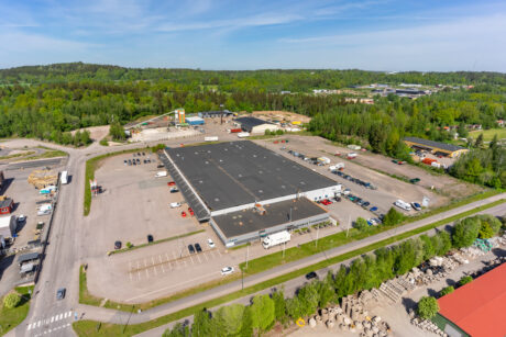 Flygfoto över en stor lagerbyggnad med grått tak, omgiven av en parkeringsplats, lastbilar, bilar och grönska en klar dag.