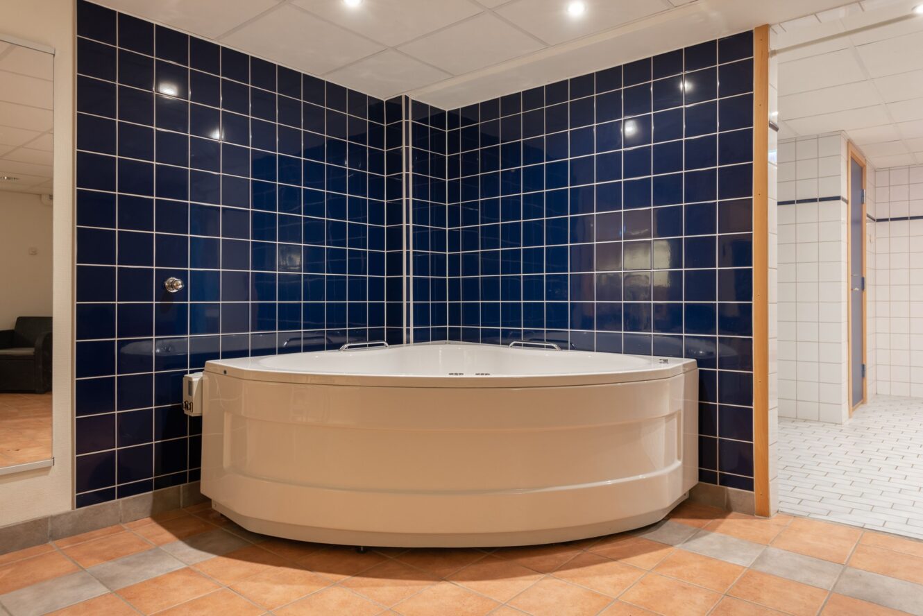 Ett vitt hörnbadkar står mot mörkblå kaklade väggar i ett badrum med en blandning av solbruna och vita golvplattor.