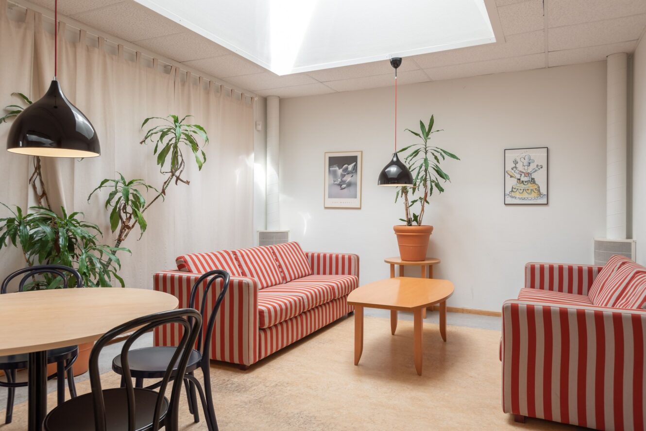 En ljus lounge med två rödvitrandiga soffor, ett soffbord i trä, ett runt matbord med stolar, växter och inramade tavlor på vita väggar.