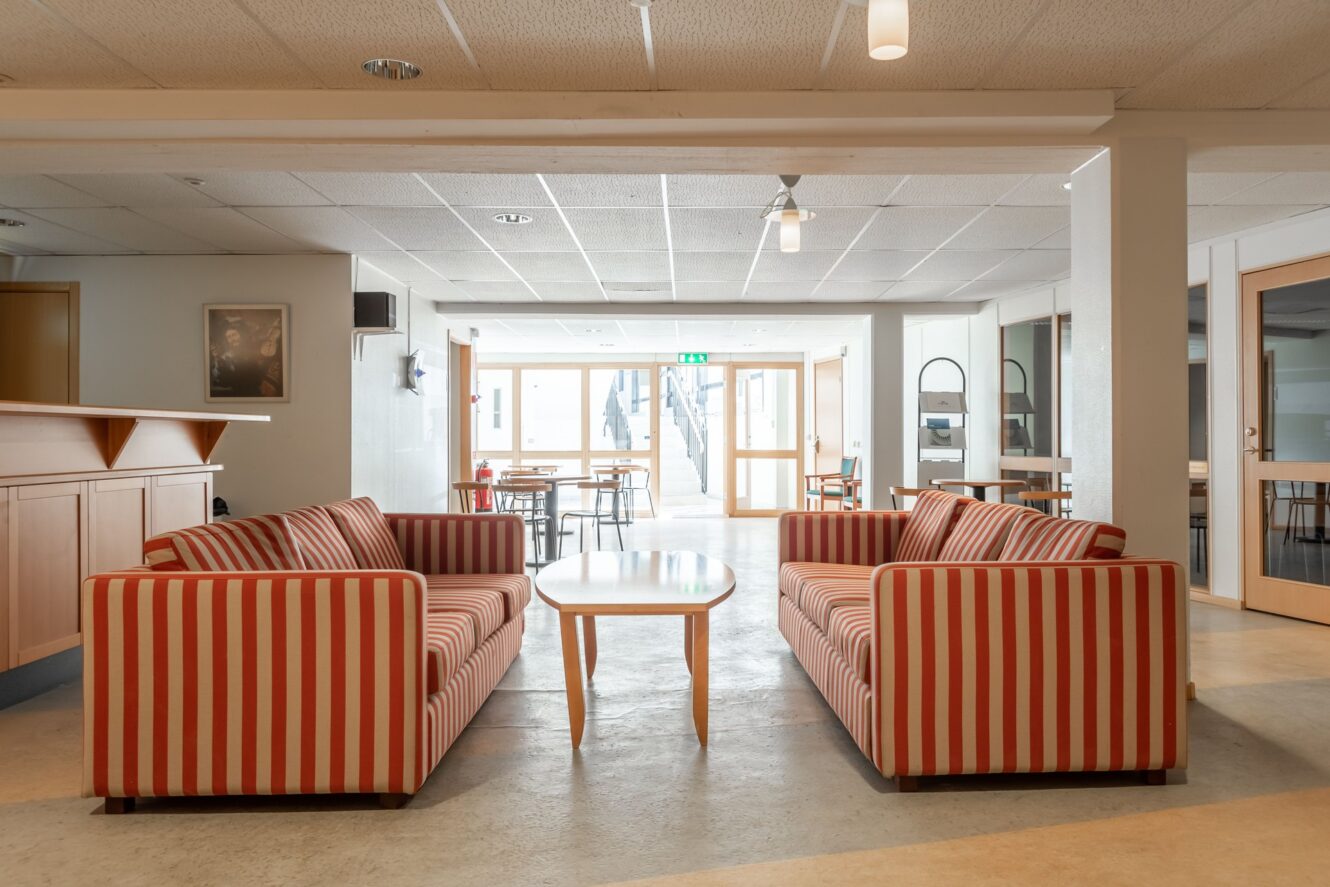 Två röd- och vitrandiga soffor står mot varandra med ett soffbord i trä mellan dem i ett ljust, modernt gemensamt utrymme med bord och stolar i bakgrunden.