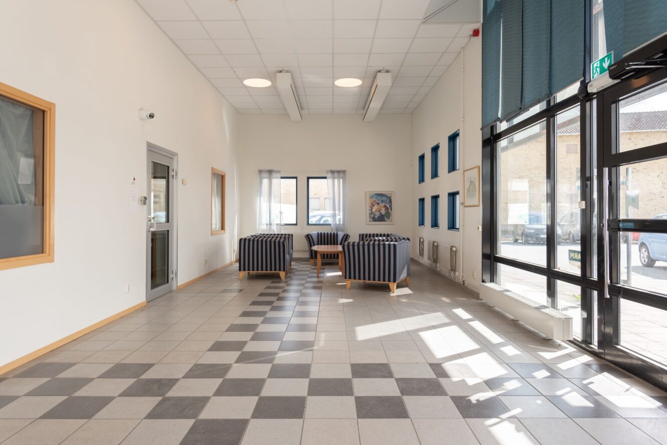 Rymligt, ljust rum med stora fönster, klinkergolv och en sittgrupp med randiga fåtöljer och ett litet bord nära den bakre väggen.