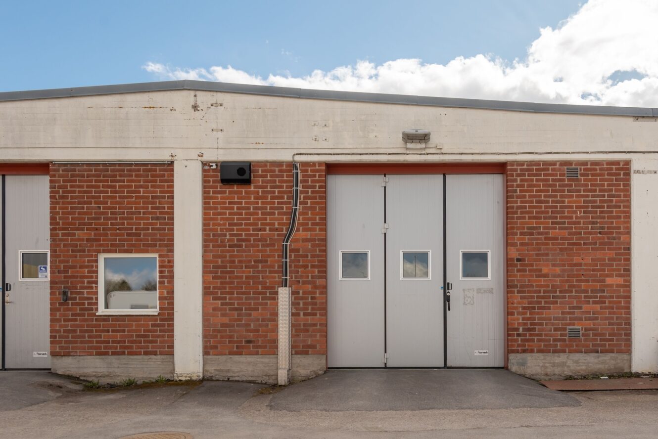 En industribyggnad i tegel med två grå garageportar, ett litet fönster och en uppfart i betong under en delvis molnig himmel.