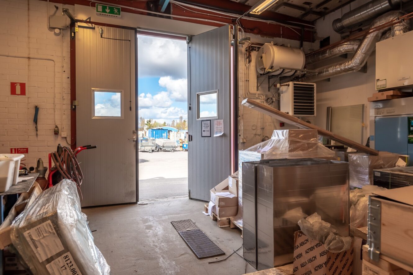 Öppna dubbeldörrar i ett industriellt förråd leder till ett utomhusområde med blå himmel och moln; olika utrustning och lådor är utspridda inuti.