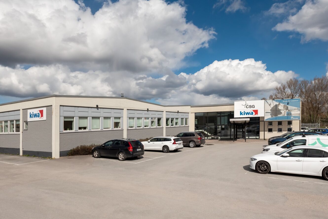 En modern affärsbyggnad med logotyperna Kiwa och CSG, flera parkerade bilar framför och en delvis molnig himmel ovanför.