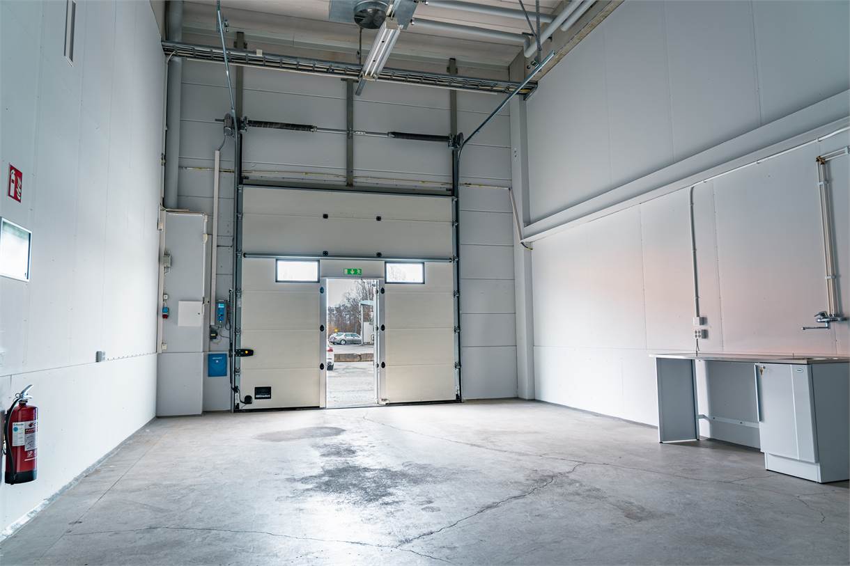 Inredning i ett tomt industrigarage med betonggolv, högt i tak, stor öppen dörr, synlig brandsläckare och en liten diskho med skåp till höger.