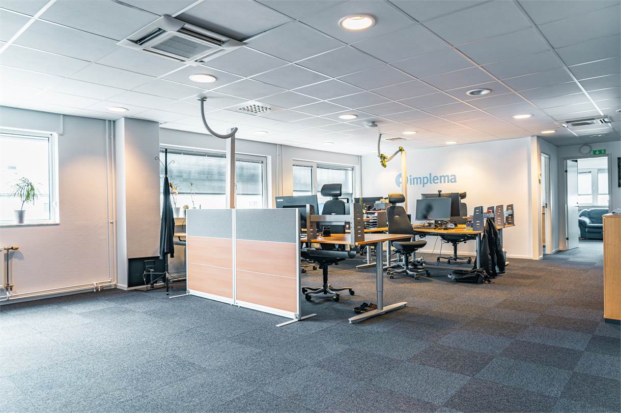 Modernt kontorslandskap med flera tomma skrivbord, stolar, datorskärmar och stora fönster som släpper in naturligt ljus. Ordet Implema står på den bakre väggen.