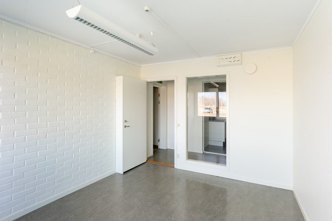Tomt, omöblerat vitt rum med en accentvägg i tegel, öppen dörr, glasfönster till ett annat rum och lysrörsarmatur i taket.