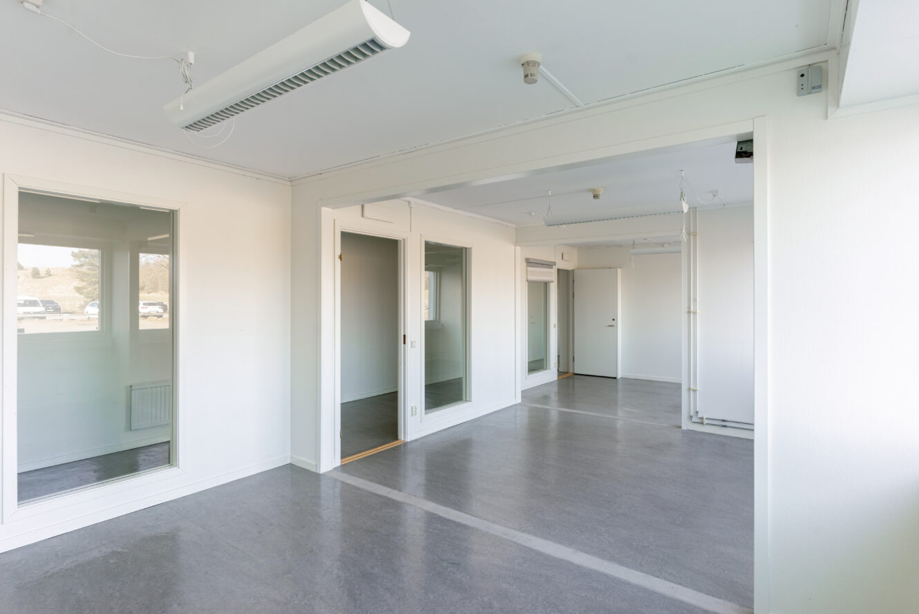 Tomt kontorsutrymme med vita väggar, stora fönster, glaspartier, grått golv och taklampor.