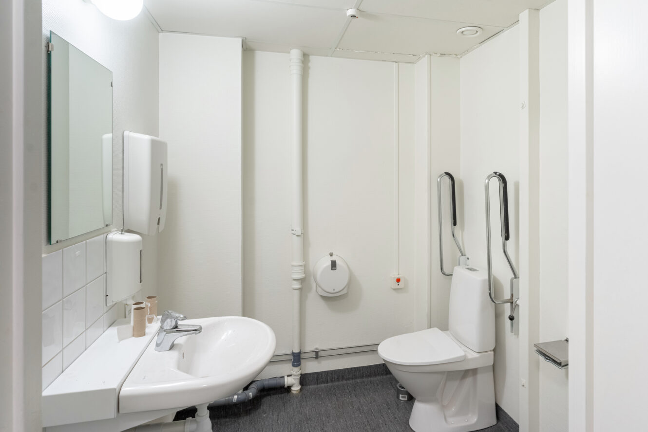 Tillgänglig offentlig toalett med en toalett utrustad med stödhandtag, ett handfat, spegel, tvålbehållare och en handtork på en vit vägg.