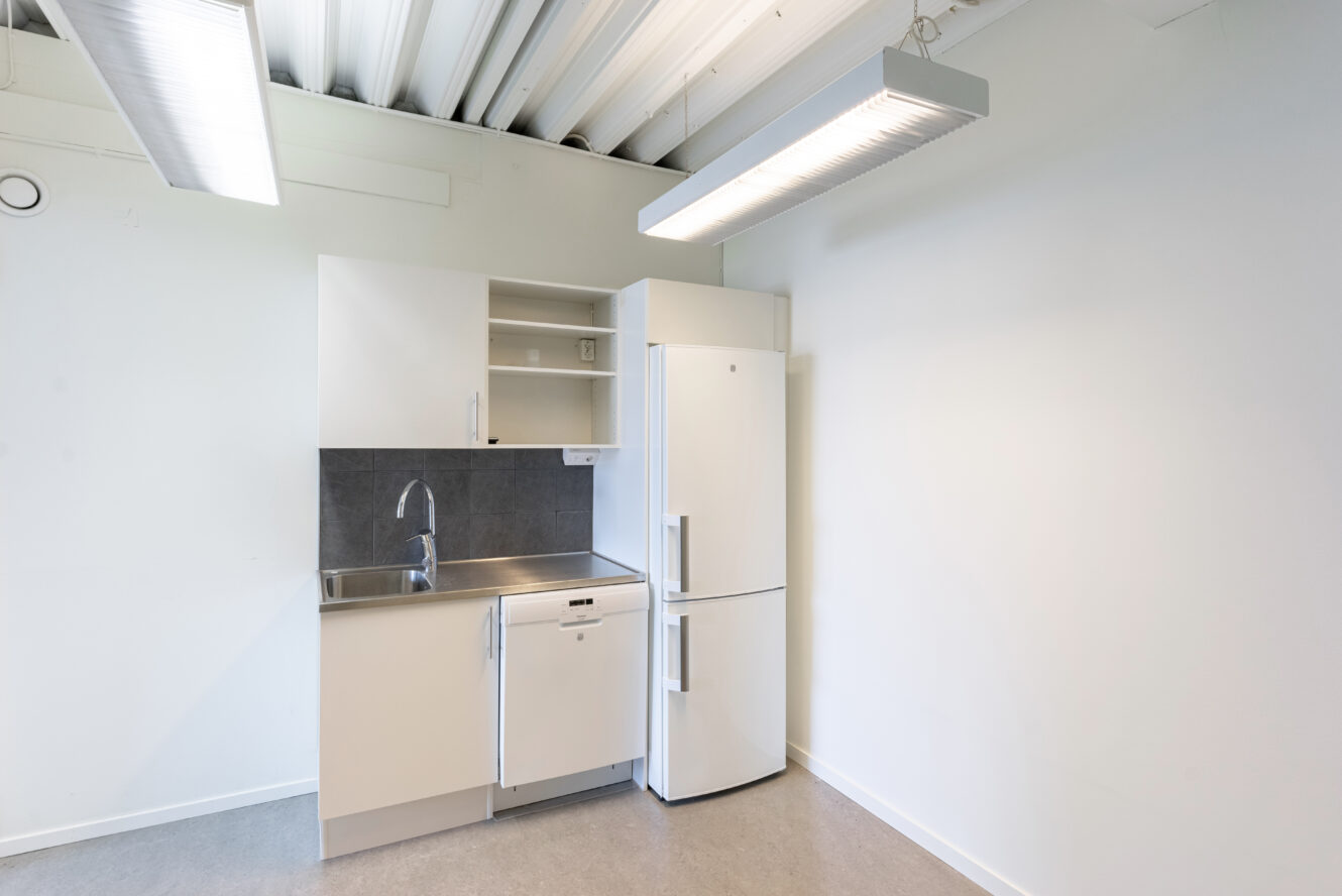 Ett litet pentry med diskho i rostfritt stål, vita skåp, diskmaskin och kylskåp, upplyst av taklampor i ett minimalistiskt rum.