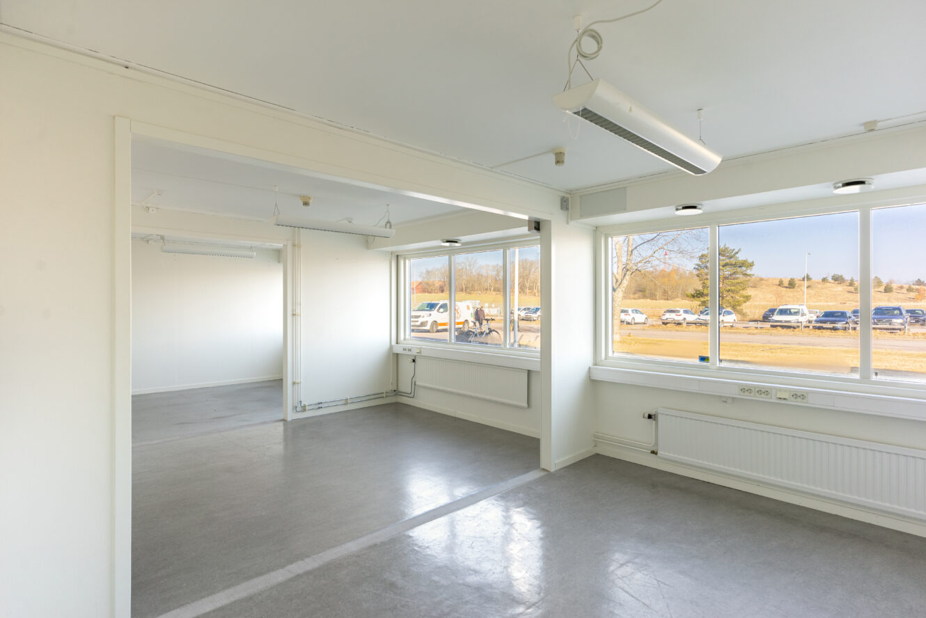 Tomt, omöblerat kontorsutrymme med vita väggar, stora fönster, grått golv och taklampor. Bilar och träd syns utanför genom fönstren.