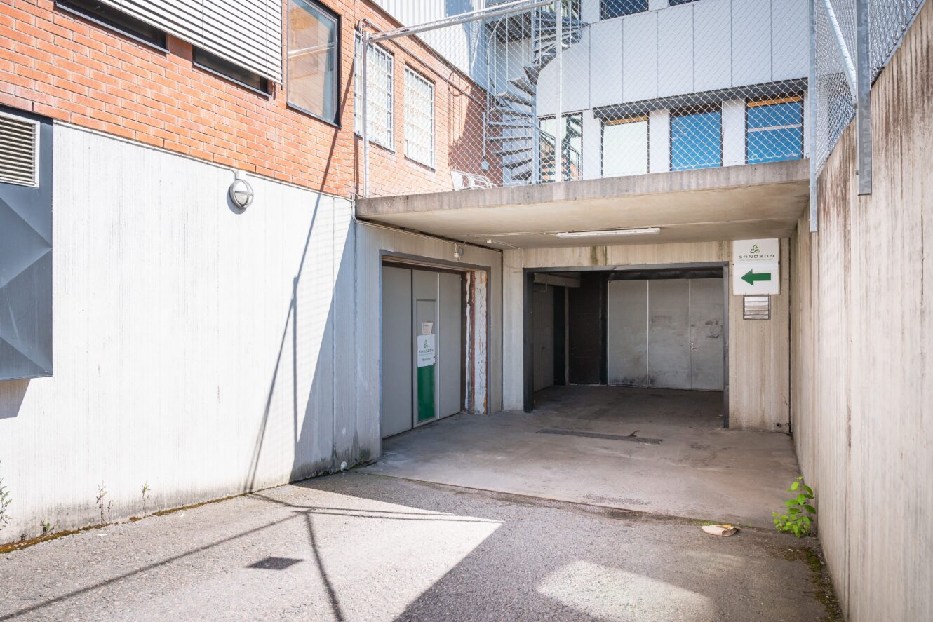 Lastkajentré i betong med garageport, taköverbyggnad och vägvisningsskyltar intill en modern industribyggnad.