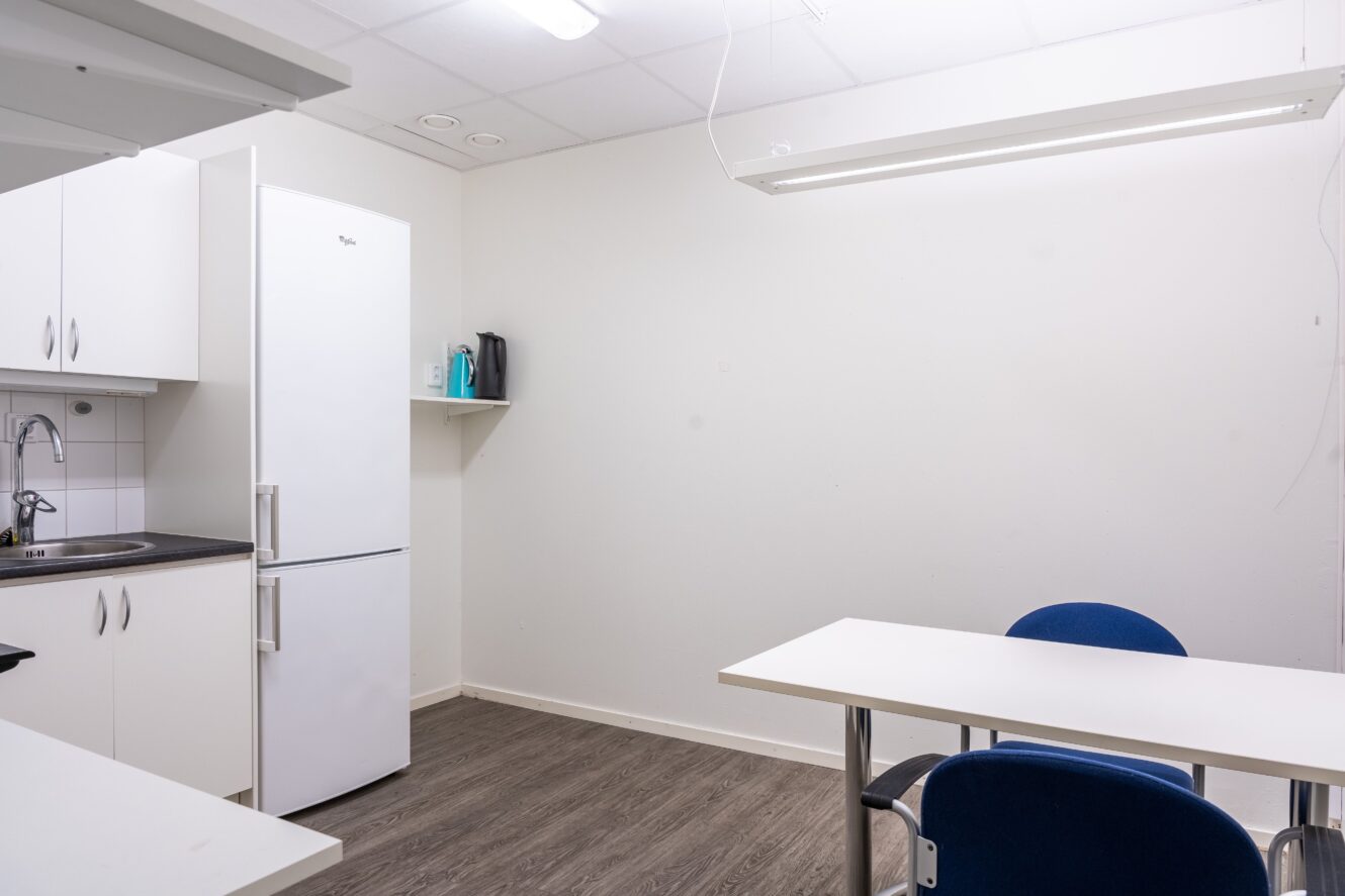 Litet fikarum med ett vitt kylskåp, diskbänk, hyllor, ett vitt bord och blå stolar under en taklampa.