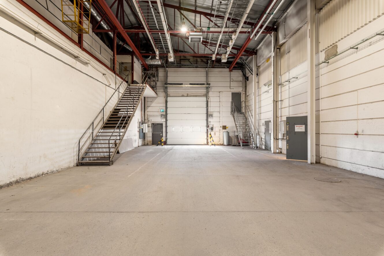 Interiör i en tom industrilagerlokal med högt i tak, metalltrappor på båda sidor, stora dörrar och synliga balkar.