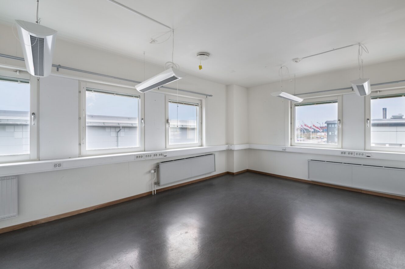 Tomt, omöblerat rum med tre stora fönster, vita väggar, mörkt golv, taklampor och synliga eluttag längs väggarna.