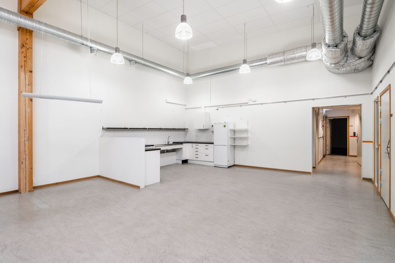 Rymligt, omöblerat rum med en liten köksdel, vita väggar, synliga kanaler, stark belysning och minimalt med hyllor.