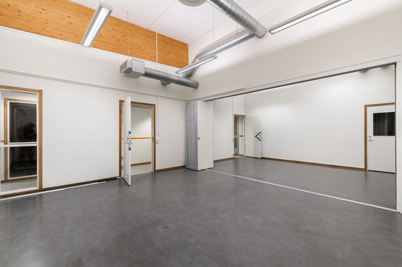 Tomt rum med grått golv, vita väggar, en stor spegel på ena väggen, kanalsystem i taket och öppna dörrar.