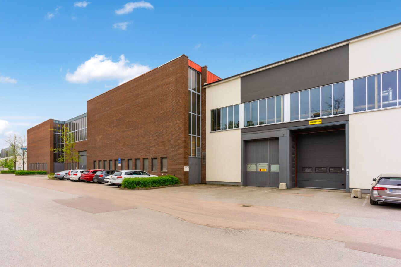 En modern industribyggnad med fasad i tegel och vitt, stora fönster, en lastkaj och flera parkerade bilar längs gatan.