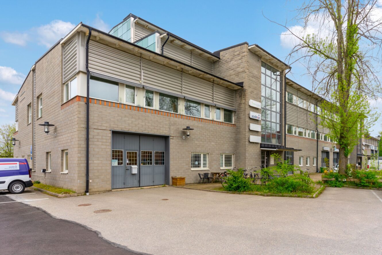 En modern kontorsbyggnad i två våningar med stora fönster och en ingång i glas, omgiven av träd och en parkeringsplats. En skåpbil står parkerad till vänster.