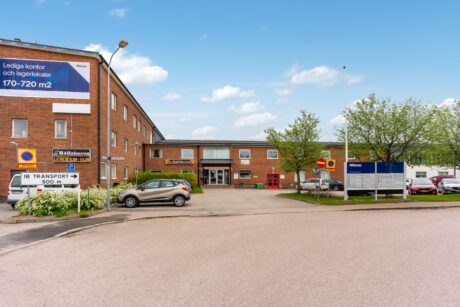 En kontorsbyggnad i tegel med skyltar på svenska, parkeringsplatser och träd framför; en stor banner annonserar lediga kontorslokaler.