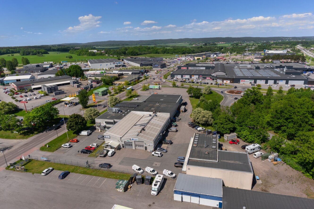 Flygfoto över ett industri- och handelsområde med lagerbyggnader, parkeringsplatser, vägar och närliggande grönska under en delvis molnig himmel.