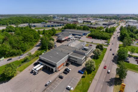 Flygfoto över ett kommersiellt och industriellt byggnadskomplex med parkeringsplats, omgivet av vägar, grönska och andra industriella strukturer under en klar himmel.