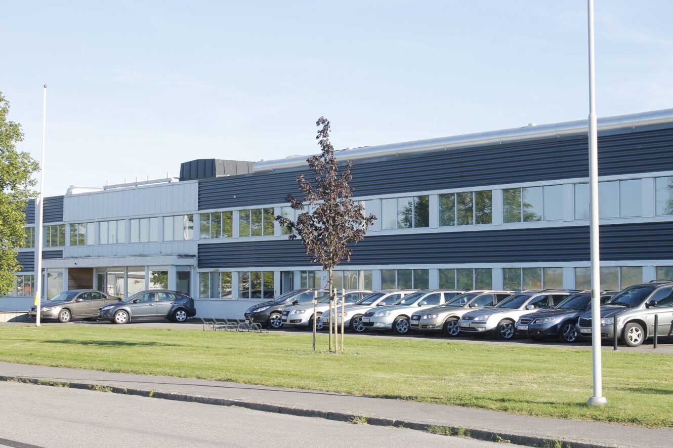 En rad bilar står parkerade framför en modern kontorsbyggnad i två våningar med stora fönster och mörk fasad.