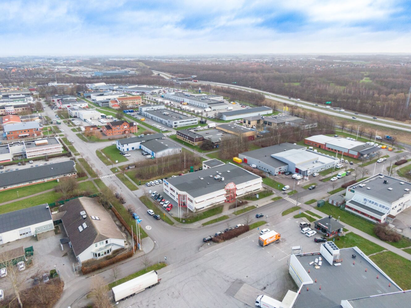 Flygfoto över ett industriområde med flera lagerlokaler, kontorsbyggnader, parkerade fordon och korsande vägar under en delvis molnig himmel.