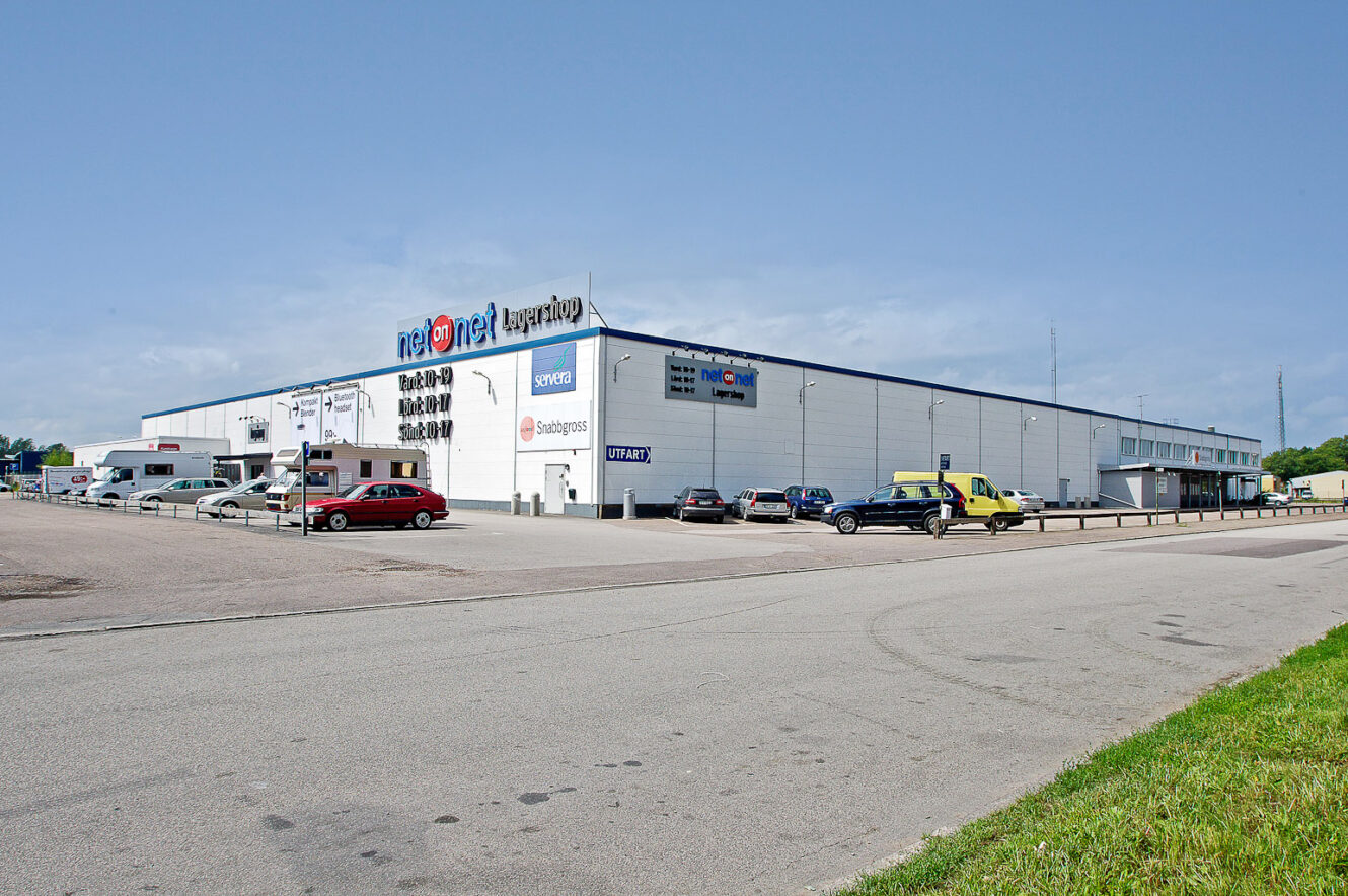 Stor vit affärsbyggnad med olika företagsskyltar på fasaden, en parkeringsplats med flera bilar och en klarblå himmel ovanför.