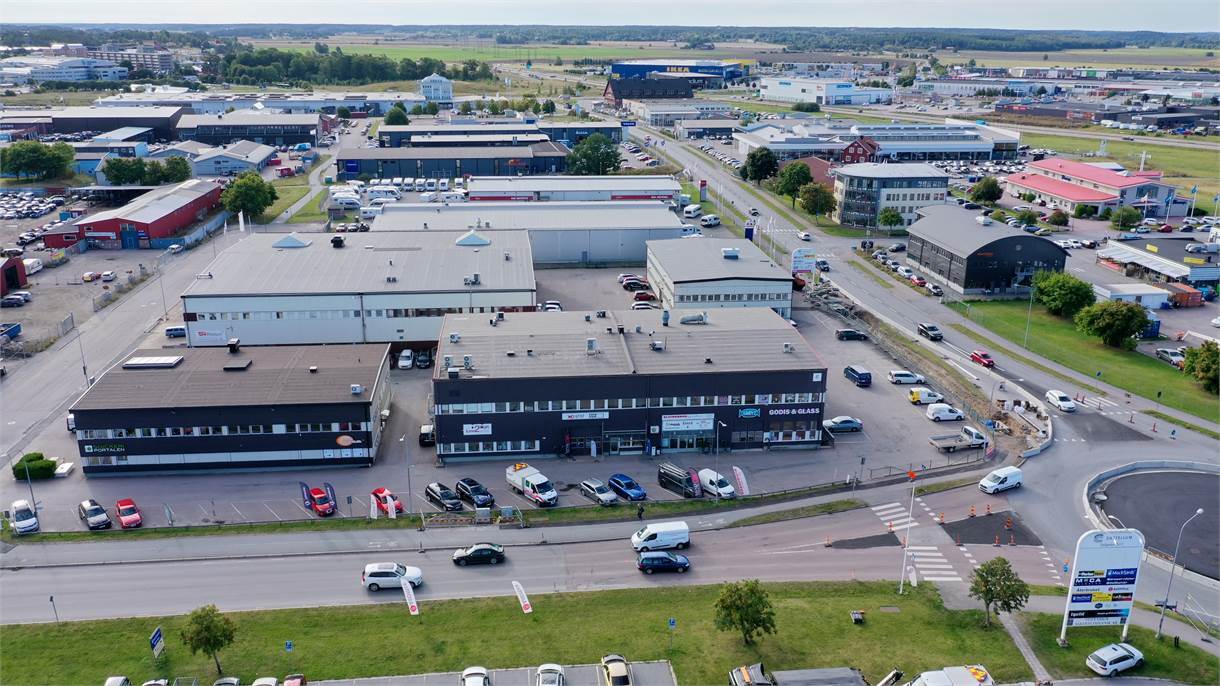 Flygfoto över ett industriområde med flera lagerlokaler, kontorsbyggnader, parkerade bilar, vägar och en rondell. Gröna fält och fler byggnader syns i bakgrunden.