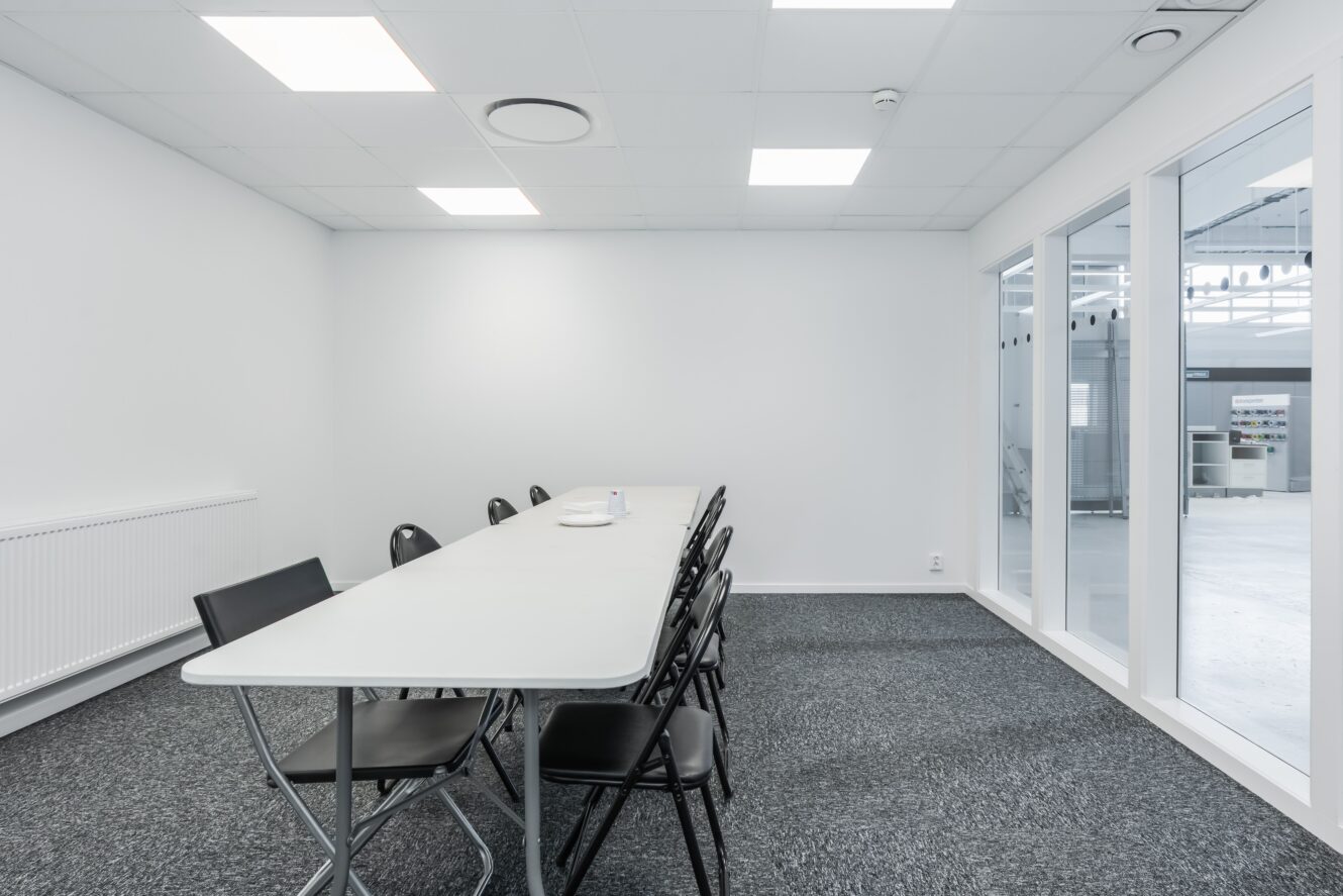 Ett enkelt konferensrum med ett långt vitt bord, åtta svarta stolar, grå heltäckningsmatta, vita väggar och stora innerfönster till höger.
