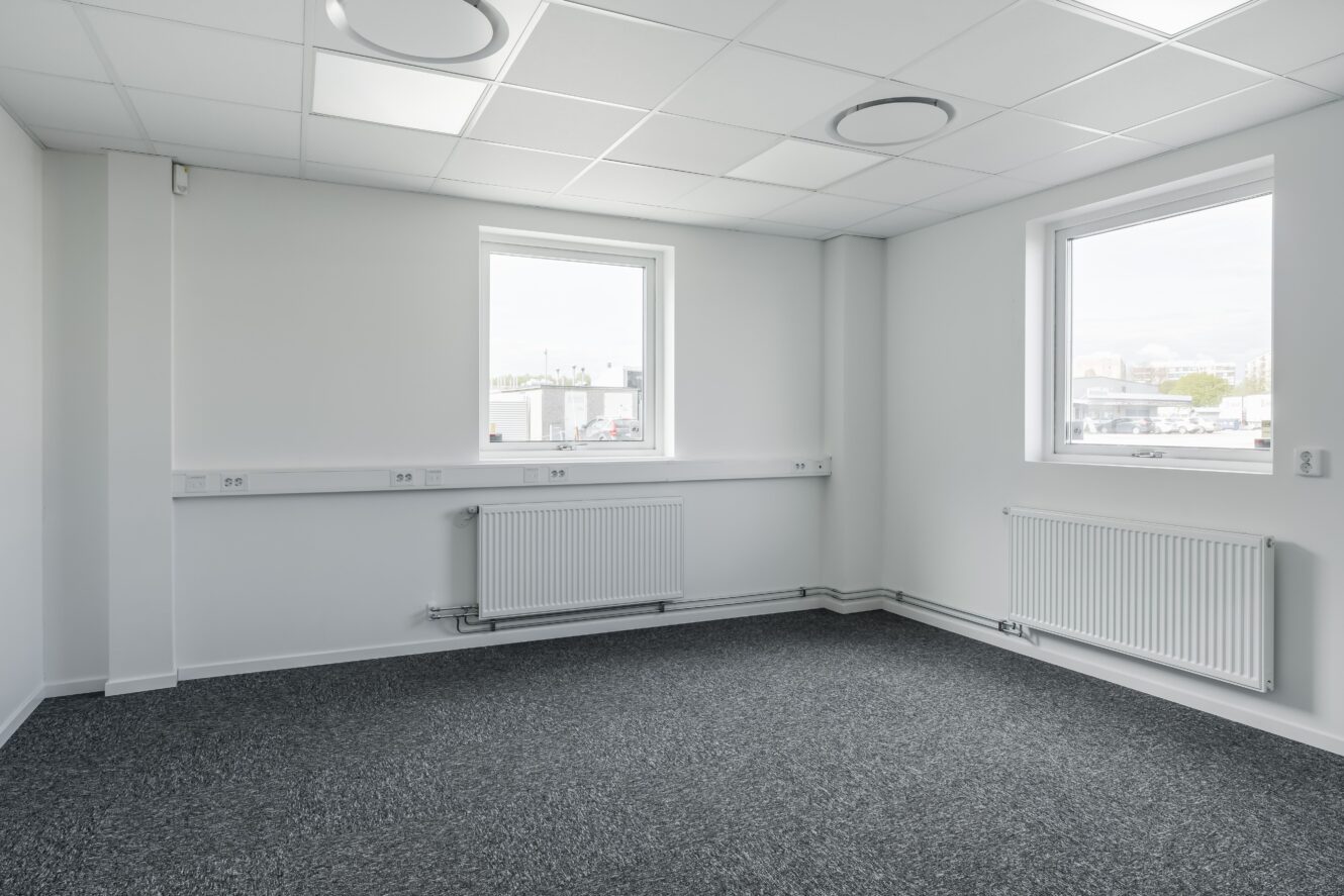 Tomt kontorsrum med vita väggar, två fönster, grå heltäckningsmatta på golvet och radiatorer under varje fönster. Taket har infällda belysningspaneler.