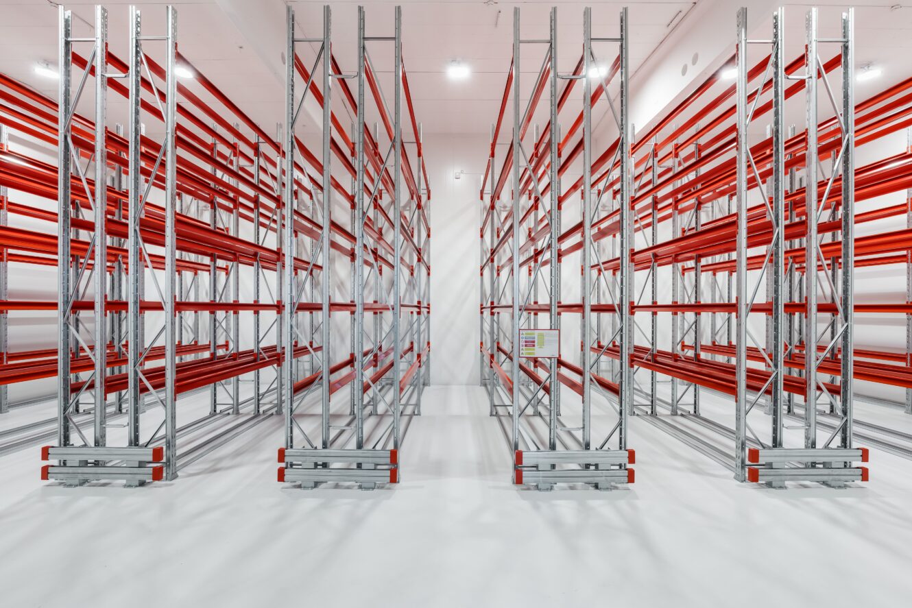 Tomma lagerhyllor av metall med röda balkar är uppställda i rader på ett rent, vitt golv under stark takbelysning.