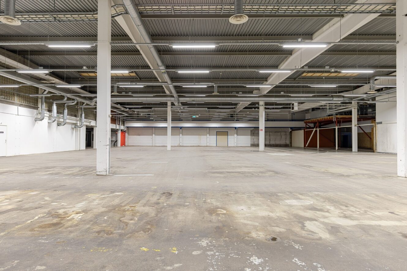 Stor tom industrilagerlokal med synliga rör i taket, betonggolv, vita väggar och lysrörsbelysning.