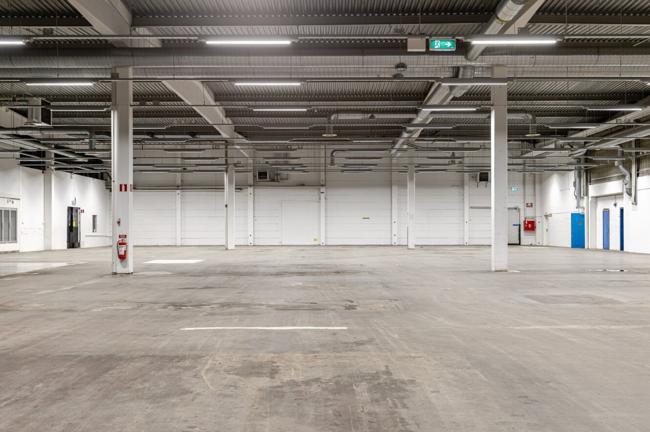 Ett tomt industrilager med betonggolv, vita väggar, synliga rör, stålbalkar och flera brandsäkerhetsutrustningar monterade på pelare och väggar.