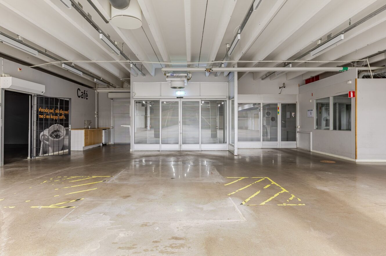 Tomt inomhusutrymme med polerat betonggolv, markerat med gul tejp; stängt café med metalljalusier och minimala möbler som syns i bakgrunden.