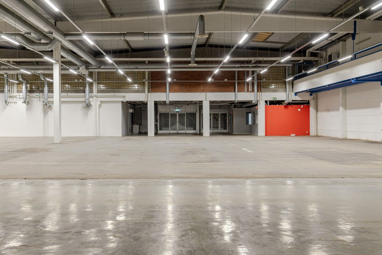 Interiör i ett stort, tomt industrilager med polerade betonggolv, takbelysning, synliga rör och en röd accentvägg till höger.