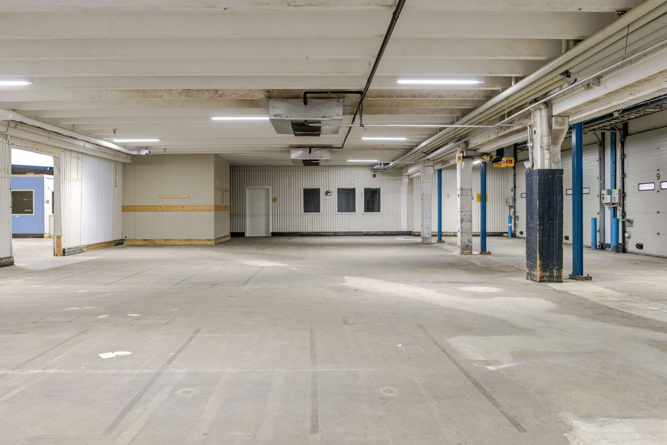 Tomt industrilager med betonggolv, rörledningar, lysrör och flera blå lastkajsdörrar längs ena väggen.