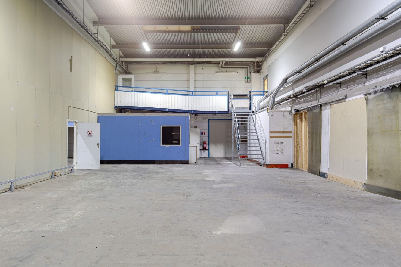 Stort tomt industrilager med betonggolv, högt i tak, blå kontorsstruktur, metalltrappa och lysrörsbelysning.