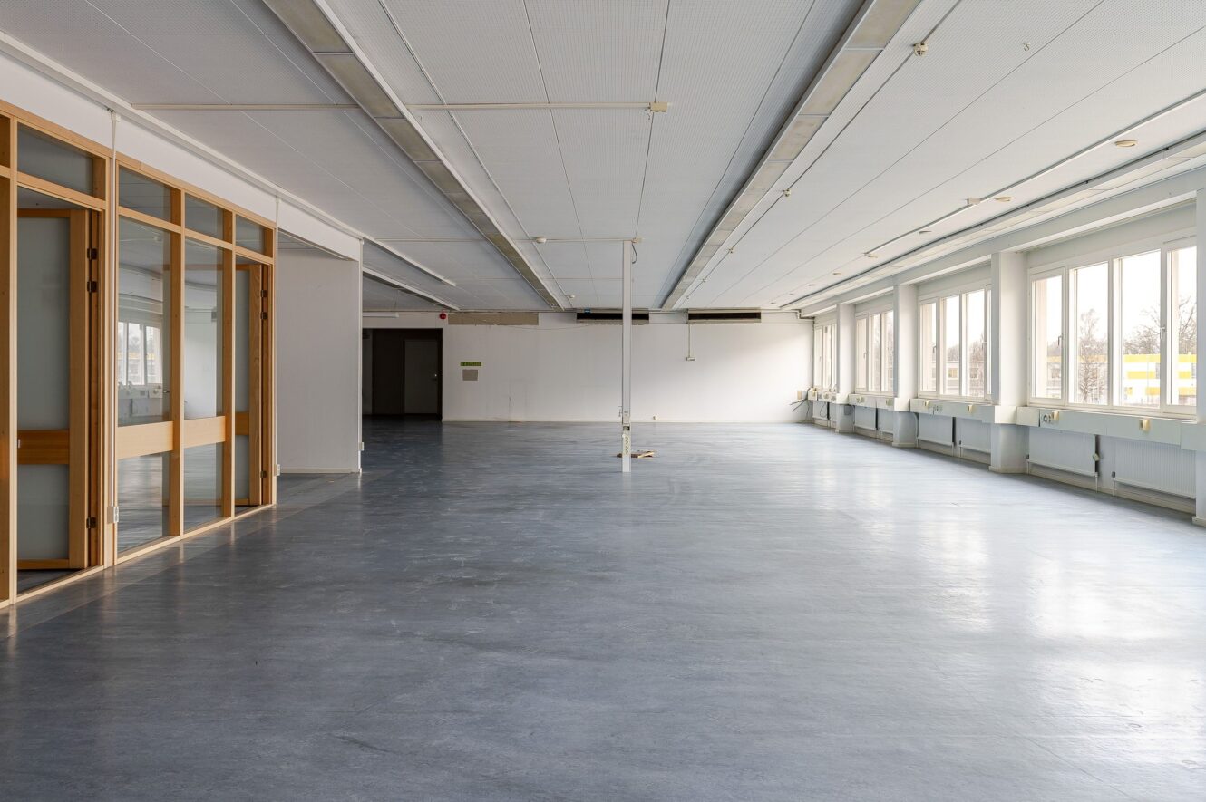 Stort, tomt kontorsutrymme med grått golv, vita väggar, glaspartier till vänster och stora fönster längs höger sida som släpper in naturligt ljus.