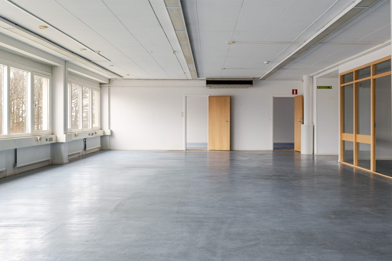 Tomt kontorsutrymme med stora fönster till vänster, släta väggar, grått golv och trädörrar på andra sidan. En dörröppning leder till ett annat rum.