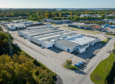En stor industrihall med vitt tak omges av vägar, parkeringsplatser och grönska i en industripark under en klar himmel.