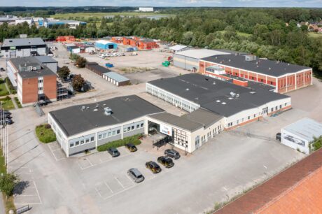 Flygfoto över ett industrikomplex med flera byggnader, parkerade bilar och containrar, omgivet av träd och öppen mark.