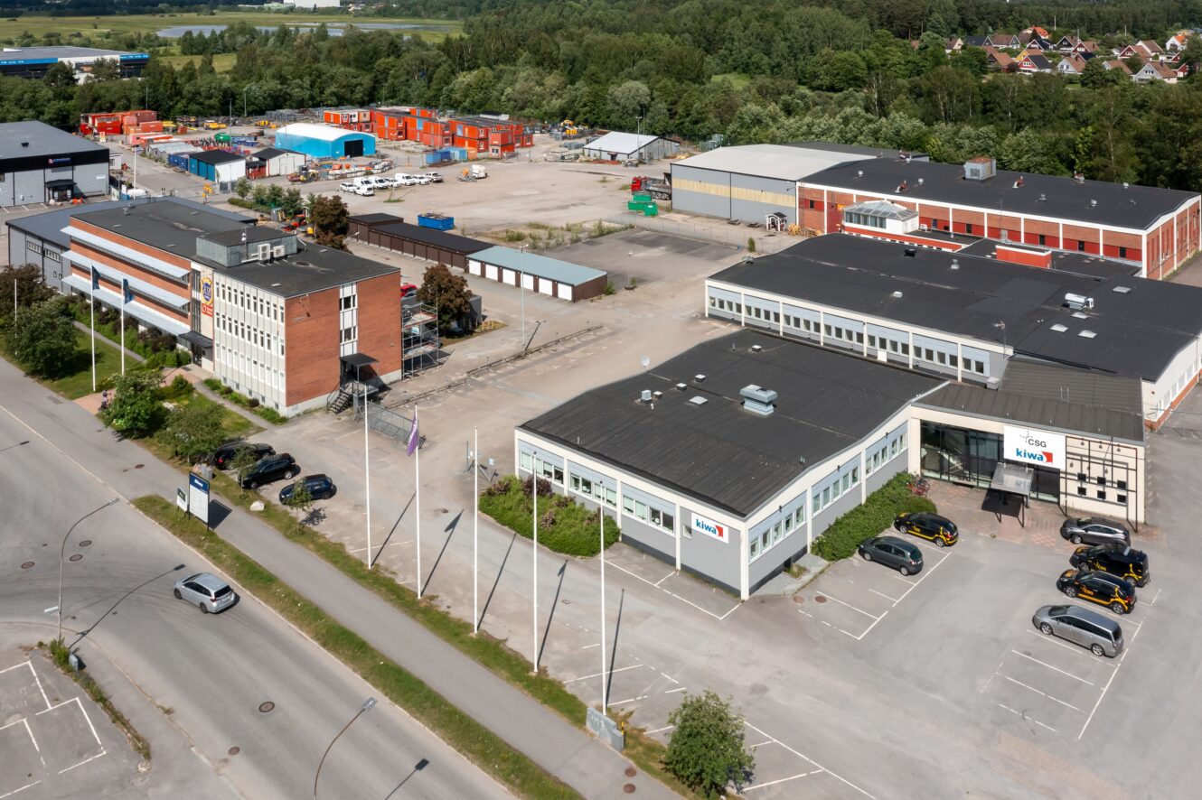 Flygfoto över ett industriområde med kontorsbyggnader, parkeringsplatser, flera bilar och byggmaterial; träd och bostadshus i bakgrunden.