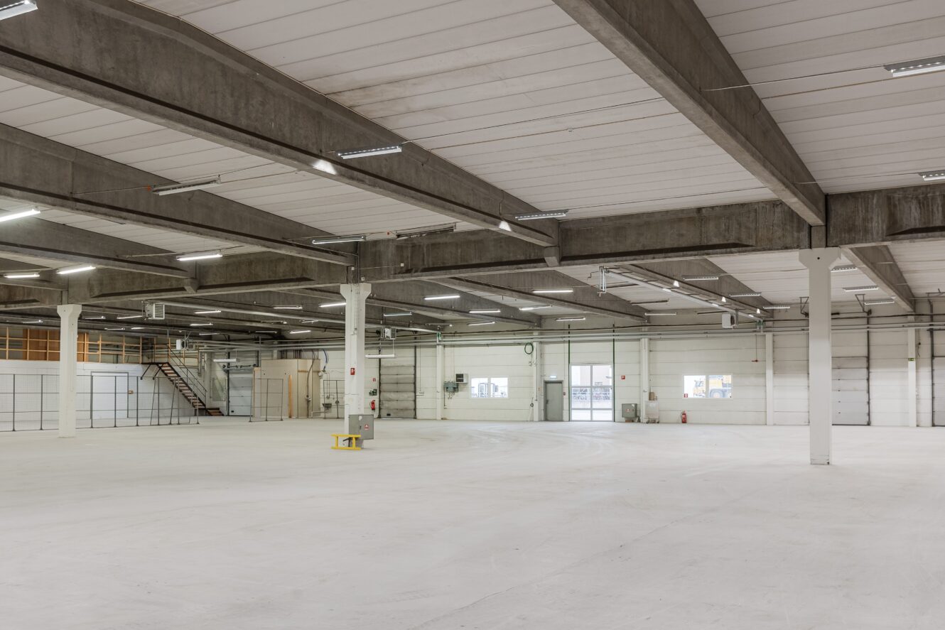 Stort, tomt industrilager med betongbjälkar, vita väggar, taklampor och ett litet gult bord i mitten. Trappa och entresol syns i bakgrunden.