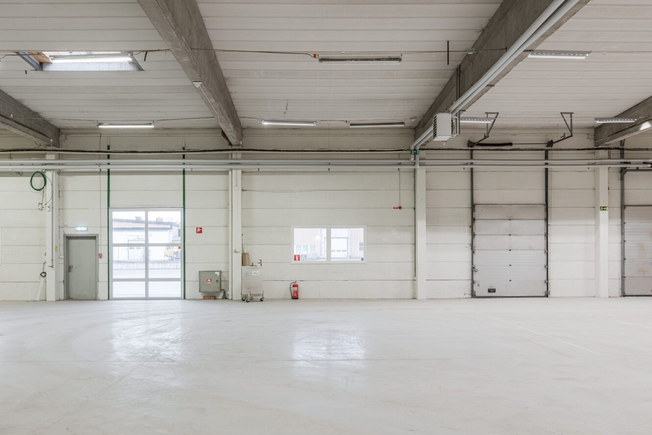En tom lagerlokal med vita väggar, högt i tak, lysrör, fönster, en dörr och en stängd garageport.