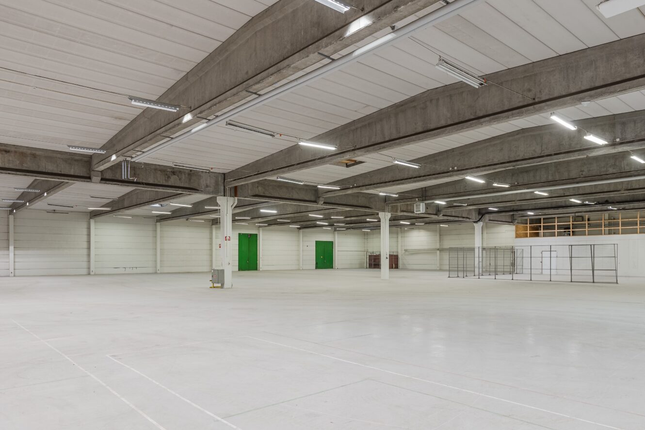 Tomt industrilager med betonggolv, synliga takbjälkar, lysrör, gröna dörrar och några förvaringsställ i metall mot väggen.